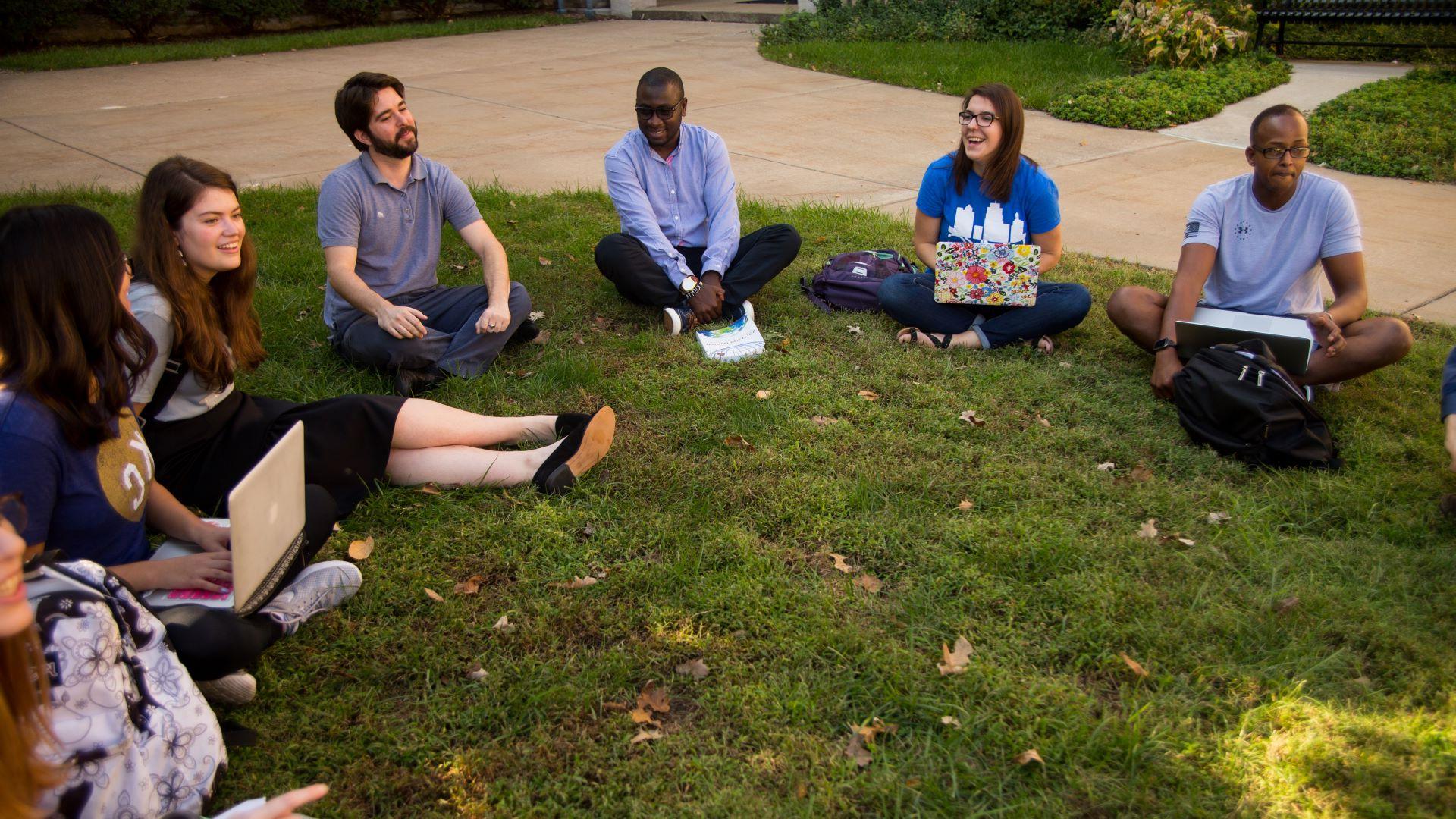 学生们在校园草坪上围成一圈坐着. 他们正在和政治学教授进行讨论. 有些人把打开的笔记本电脑放在腿上.